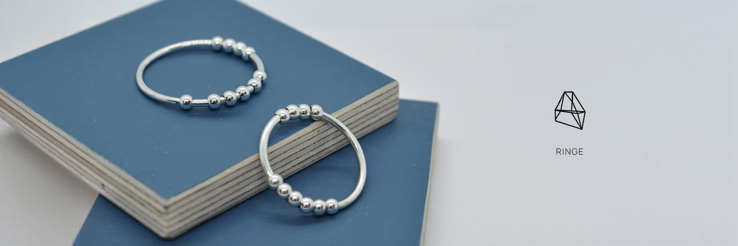 DOPPELLUDWIG © 2026 Zwei DOPPELLUDWIG Ringe mit kleinen Silberperlen ruhen auf gestapelten blauen und weißen quadratischen Platten. Auf der rechten Seite sind ein minimalistisches geometrisches Ringsymbol und das Wort "RINGS" auf einem hellen Hintergrund zu sehen.