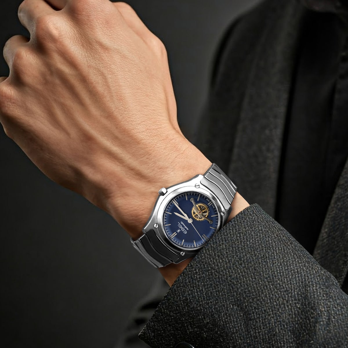 EBEL © 2026 Nahaufnahme des Handgelenks einer Person, die eine silberne Ebel-Metalluhr mit blauem Zifferblatt und sichtbarem mechanischem Uhrwerk trägt, gekleidet in eine dunkle, strukturierte Anzugsjacke vor einem dunklen Hintergrund.