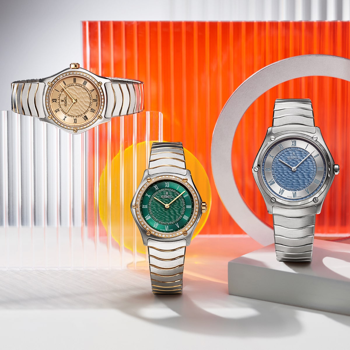 EBEL © 2026 Drei luxuriöse Ebel-Armbanduhren mit Metallbändern werden vor einem modernen Hintergrund aus orangefarbenen, weißen und gelben geometrischen Formen präsentiert. Die Zifferblätter leuchten in Gold, Grün und Blau.