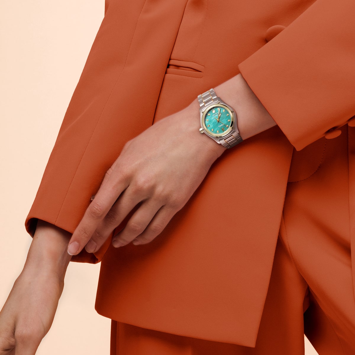 Eine Person in einem orangefarbenen Anzug trägt am linken Handgelenk eine silberne Armbanduhr mit türkisfarbenem Zifferblatt vor einem hellbeigen Hintergrund.
