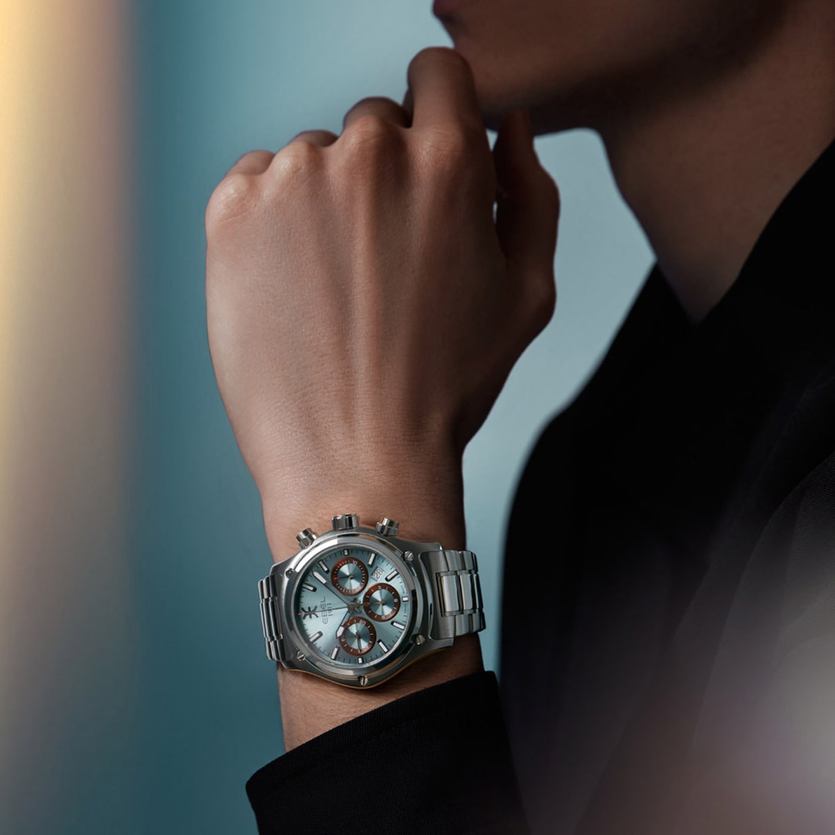 Eine Person, die eine silberne Ebel-Armbanduhr mit einem großen, runden Zifferblatt und mehreren Ziffernblättern trägt, stützt ihr Kinn auf ihre Hand vor einem sanften blauen Hintergrund. Die Person ist dunkel gekleidet.