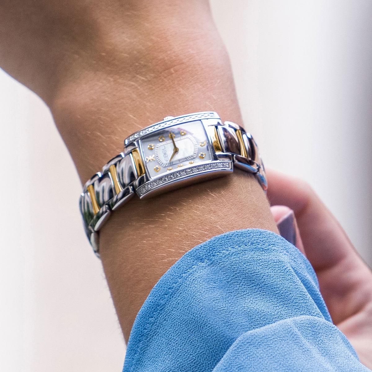 Nahaufnahme eines Handgelenks, das die elegante Ebel Brasilia trägt, eine rechteckige Uhr in Silber und Gold mit diamantbesetzter Lünette und blau-goldenem Metallgliederband. Die Person ist mit einem hellblauen, langärmeligen Oberteil bekleidet.