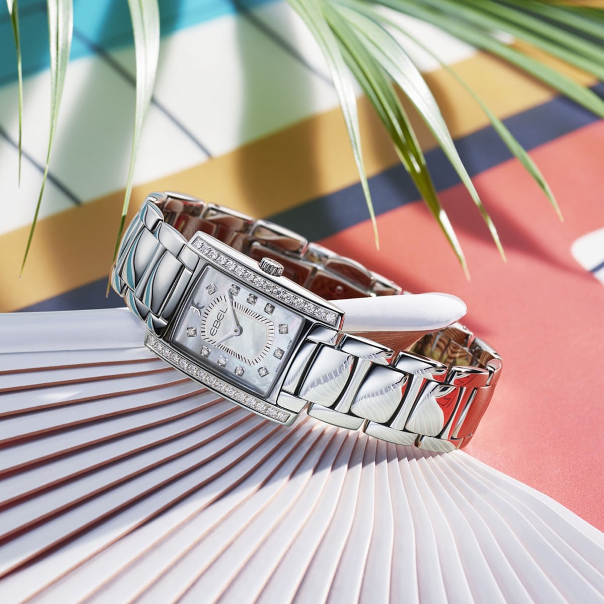 Eine silberne Ebel Brasilia-Uhr mit rechteckigem Zifferblatt und Diamantakzenten ruht auf einem gefalteten weißen Fächer mit bunten geometrischen Mustern und Palmenblättern im verschwommenen Hintergrund.