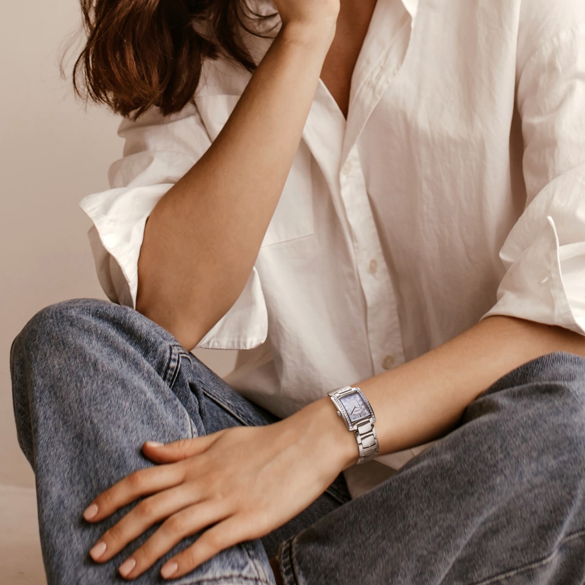 Eine Person, die ein weißes Button-up-Hemd und blaue Jeans trägt, sitzt im Schneidersitz, stützt einen Arm auf das Knie und den anderen auf den Schoß und präsentiert eine elegante silberne Ebel Brasilia-Armbanduhr. Ihr Gesicht ist teilweise außerhalb des Rahmens.