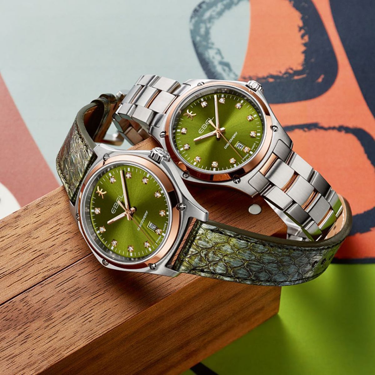 Zwei Ebel Discovery Armbanduhren mit grünen Zifferblättern und silberfarbenen Gehäusen werden ausgestellt. Eine hat ein silbernes Metallarmband, die andere ein grünes, strukturiertes Lederarmband. Beide ruhen auf einem Holzblock vor einem bunten Hintergrund.