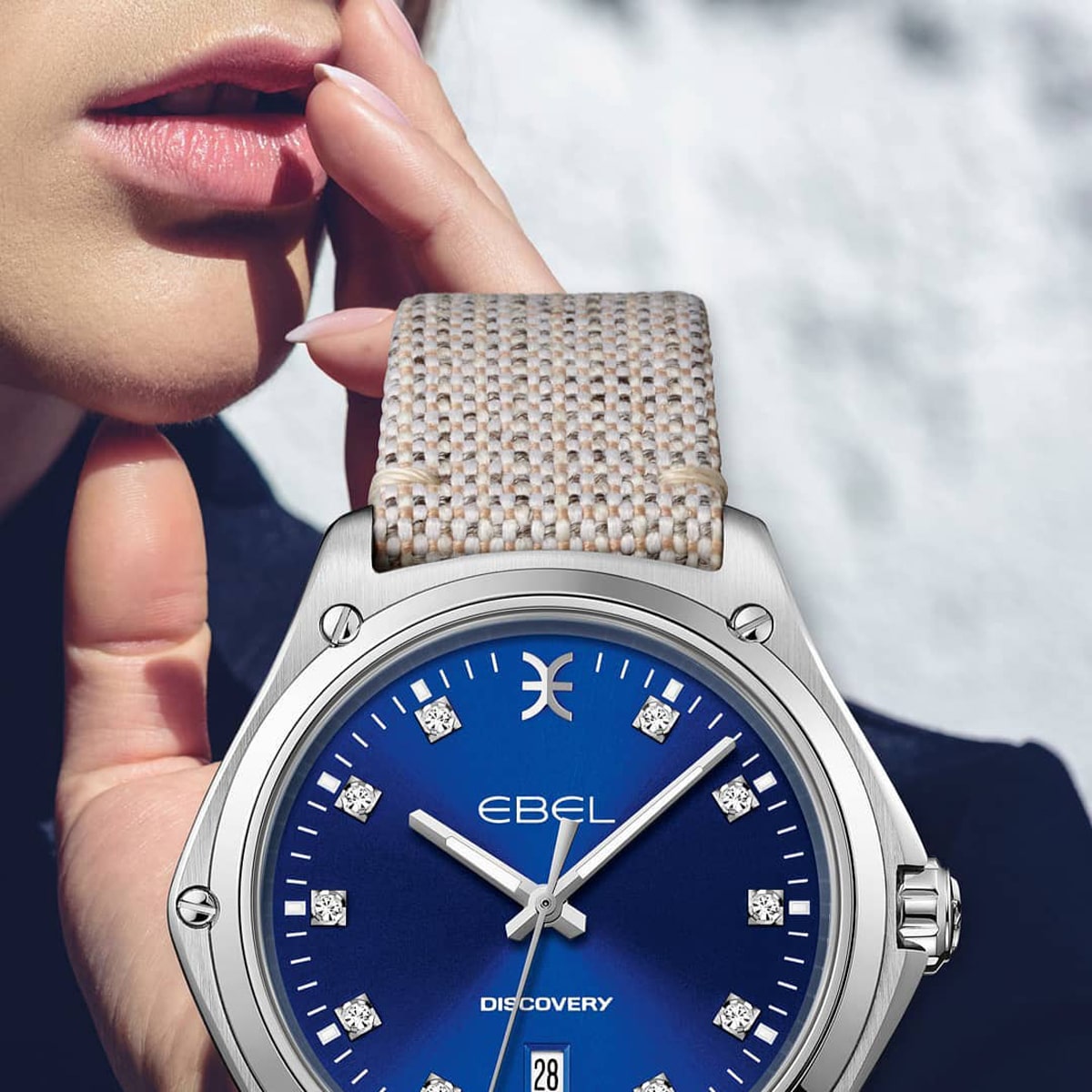 Nahaufnahme des Gesichts und der Hand einer Person in der Nähe ihrer Lippen, mit einem detaillierten Blick auf eine silberne und blaue Ebel Discovery Armbanduhr mit einem gewebten beigen Armband und Diamantindexen.