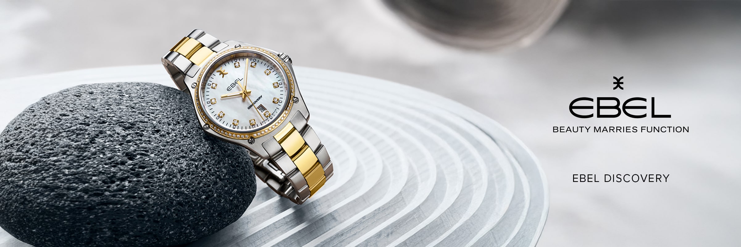 Eine goldene und silberne EBEL-Armbanduhr mit weißem Zifferblatt ruht auf einem glatten schwarzen Stein, der auf einer strukturierten Oberfläche liegt. Das EBEL Logo und die Worte "Beauty Marries Function" und "Ebel Discovery" erscheinen elegant auf der rechten Seite.