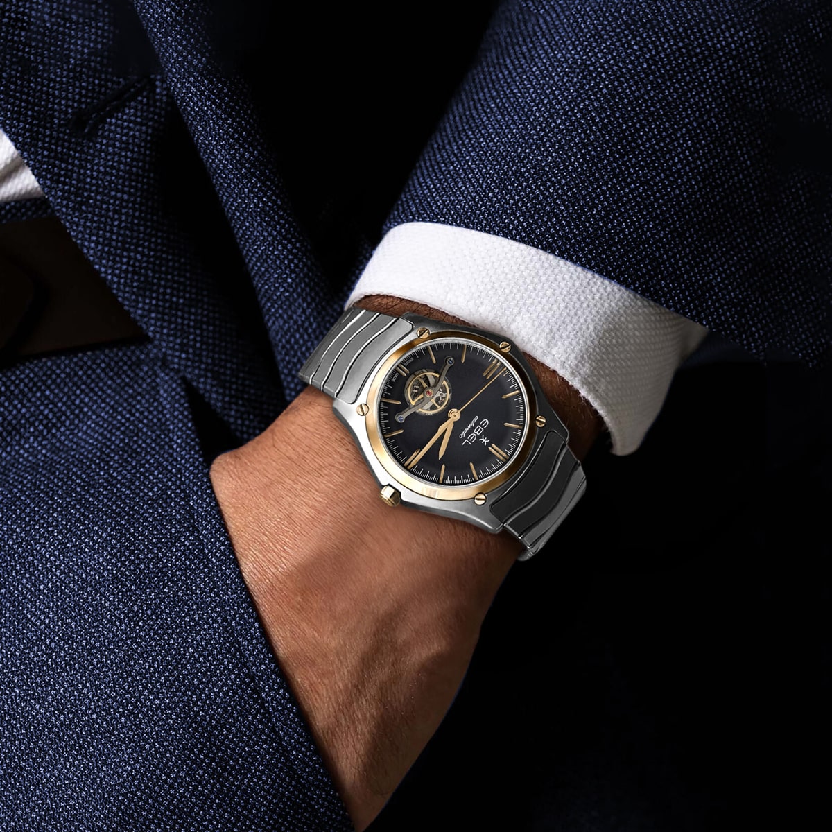 Eine Person, die einen dunkelblauen Anzug und ein weißes Hemd trägt, hat die Hand in der Tasche und präsentiert eine silberne Ebel Sport Classic Armbanduhr mit schwarzem Zifferblatt und goldenen Akzenten.