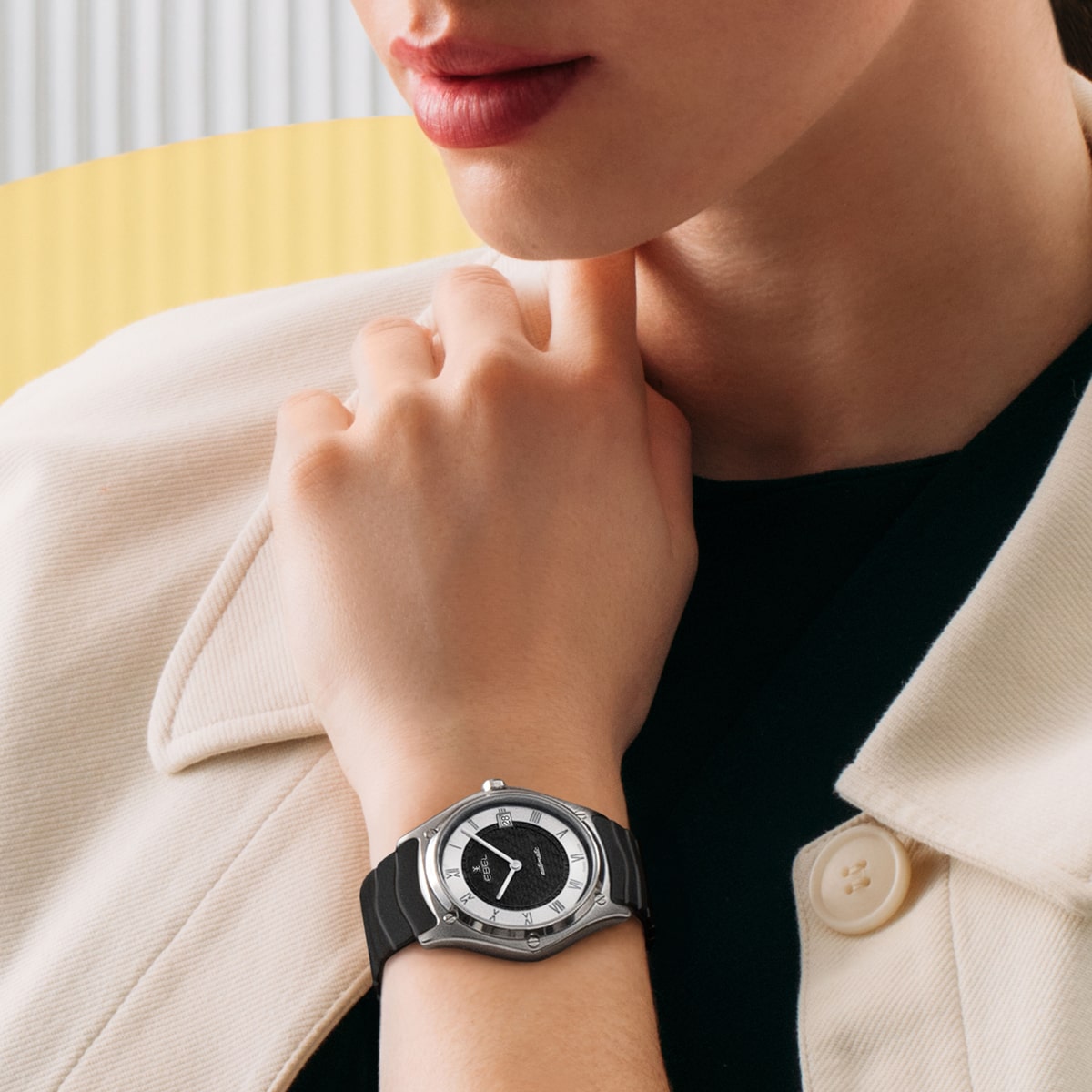 Eine Person, die eine cremefarbene Jacke und ein schwarzes Oberteil trägt, stützt ihr Kinn auf ihre Hand und präsentiert eine silber-schwarze Armbanduhr mit rundem Zifferblatt und schwarzem Armband. Der Hintergrund ist in sanften Gelb- und Weißtönen gehalten.