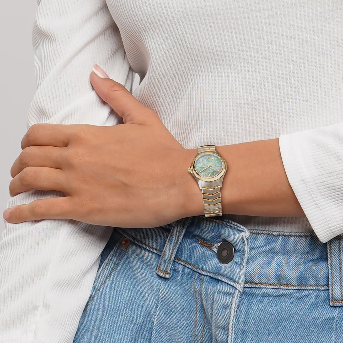 Eine Person, die eine goldene Ebel Wave Armbanduhr mit hellblauem Zifferblatt trägt, bekleidet mit einem weißen langärmeligen Rippenhemd und blauen Jeans, die einen Arm über dem Körper verschränkt.