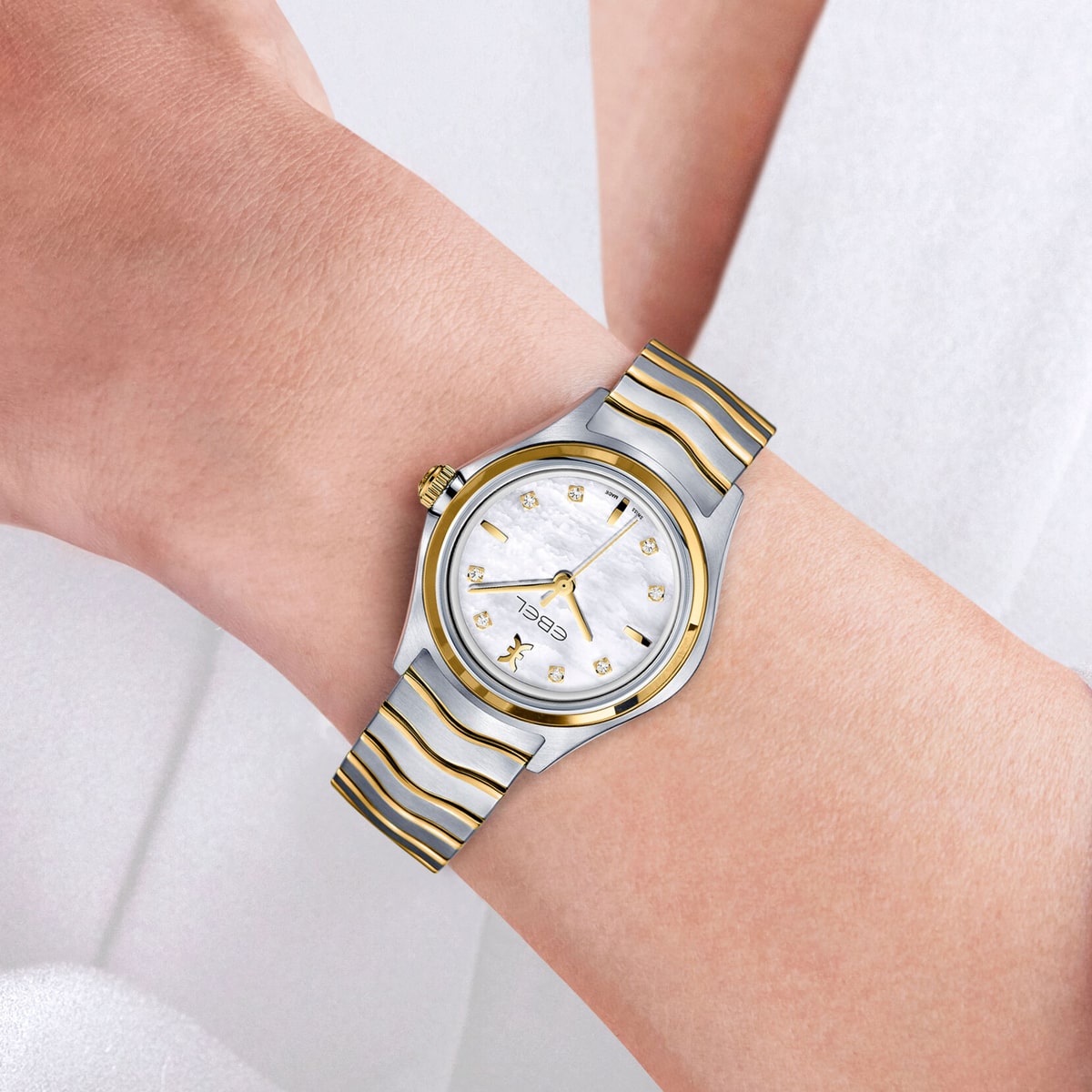 Nahaufnahme eines Handgelenks, das eine silberne und goldene Ebel Wave Uhr mit weißem, gemustertem Zifferblatt, goldenen Indexen und einer goldenen Krone trägt. Die Person ist mit heller Kleidung bekleidet.