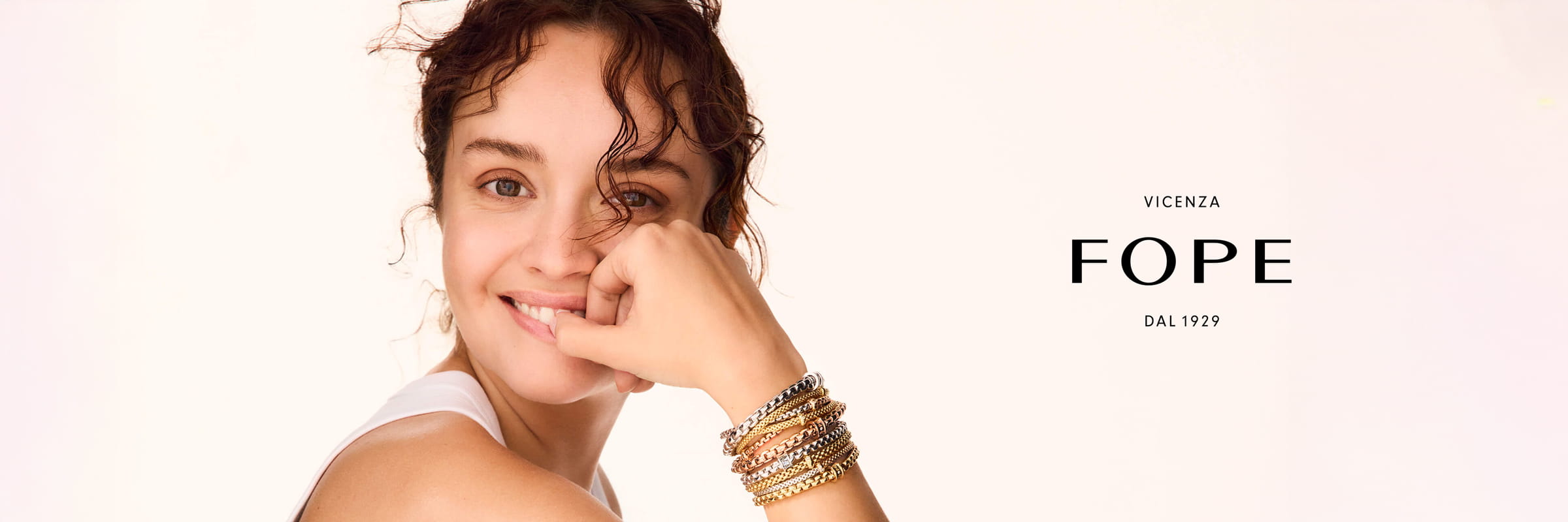FOPE © 2026 Eine Frau mit lockigem Haar lächelt in die Kamera und stützt ihr Kinn auf ihre Hand. Sie trägt mehrere goldene und silberne Armbänder von Fope. Der Text neben ihr lautet: "VICENZA FOPE DAL 1929" auf hellem Hintergrund.