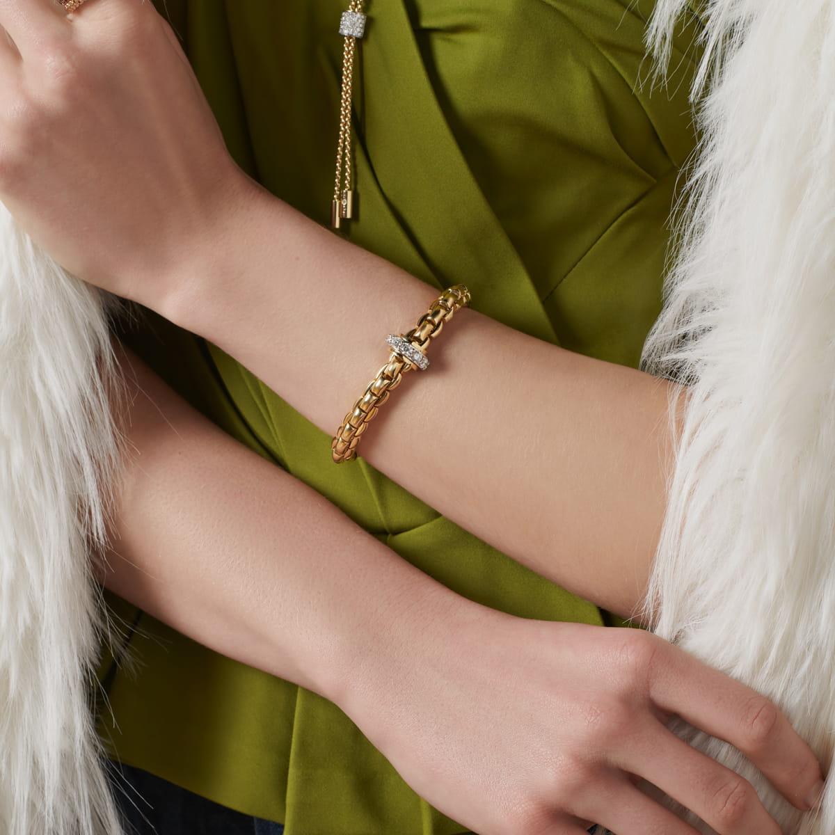 Eine Person, die ein goldenes Kettenarmband von Fope Armschmuck mit einem diamantbesetzten Muster trägt, bekleidet mit einem grünen Oberteil und in einen weißen Kunstpelz gehüllt. Ihre Arme sind verschränkt, wodurch der luxuriöse Schmuck elegant zur Geltung kommt.