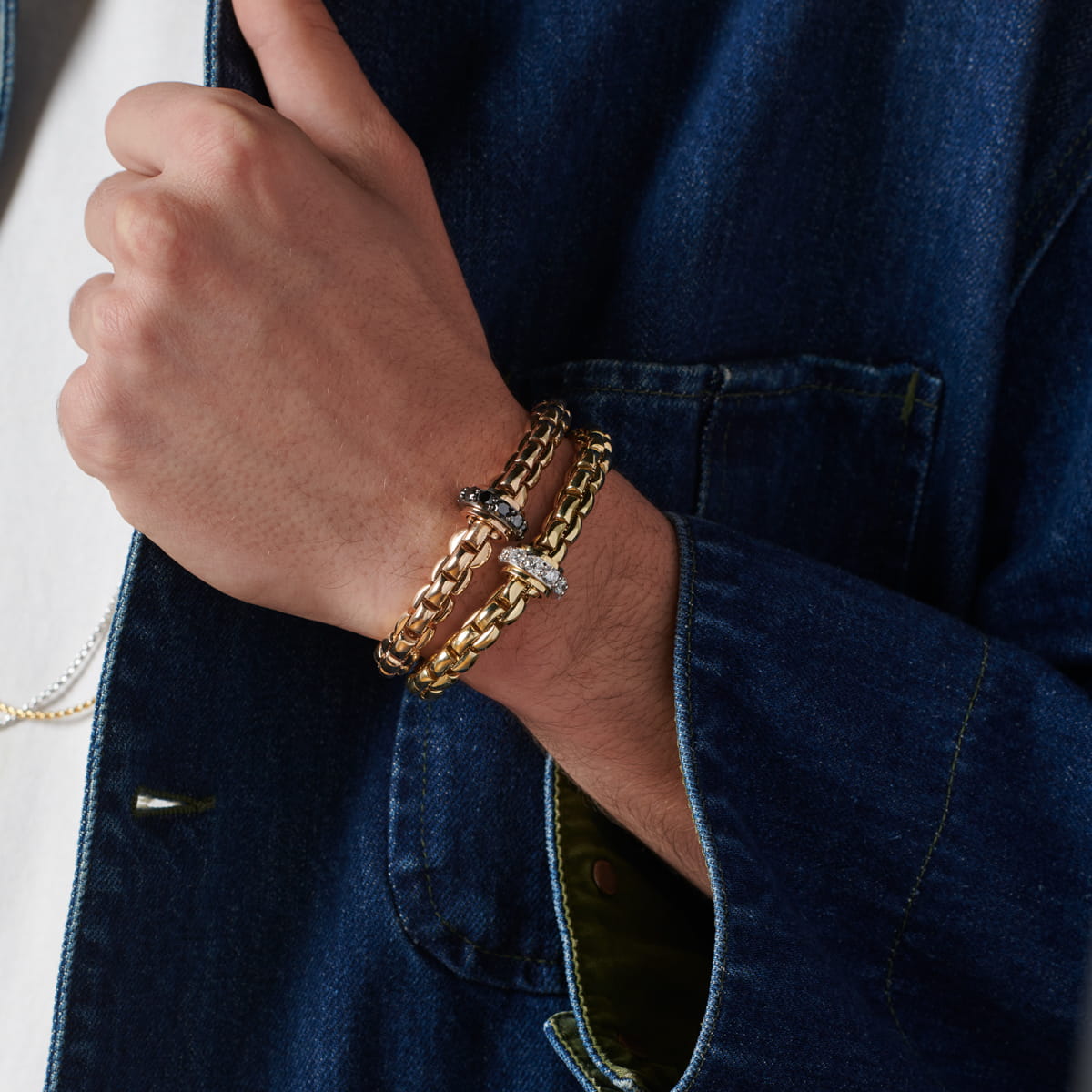 Eine Person, die eine blaue Jeansjacke und ein weißes Hemd trägt, zeigt zwei klobige Kettenarmbänder, darunter ein von Fope inspiriertes goldenes Design und eines mit dunklerem Metall, an ihrem Handgelenk. Beide Armbänder haben dekorative Verschlüsse mit funkelnden Details.