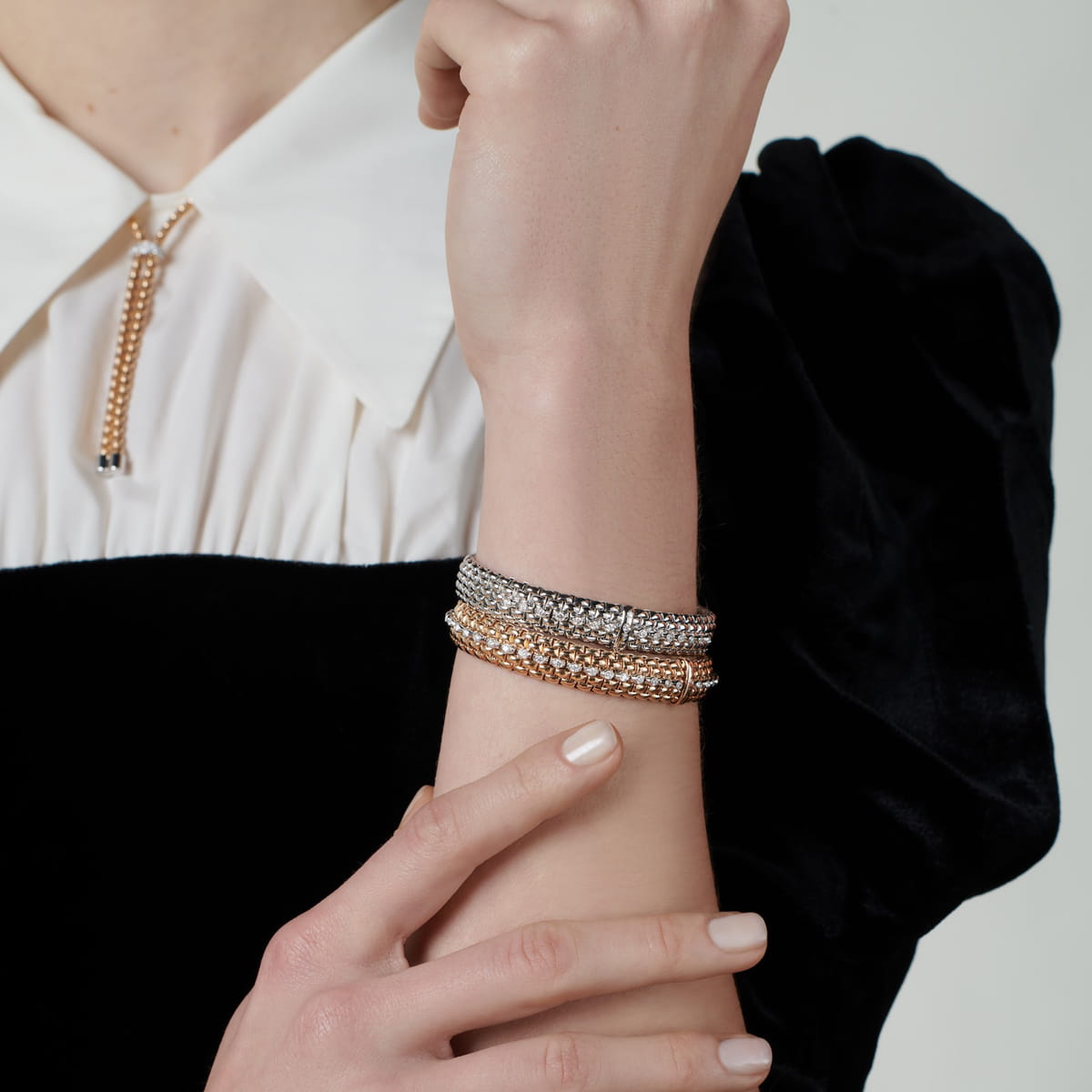 Eine Person, die ein schwarzes Samtkleid mit weißem Kragen trägt, zeigt ihr Handgelenk, das mit drei metallischen, strukturierten Fope Armschmuck Armbändern in Silber- und Goldtönen geschmückt ist. Ihre andere Hand berührt sanft das geschmückte Handgelenk.