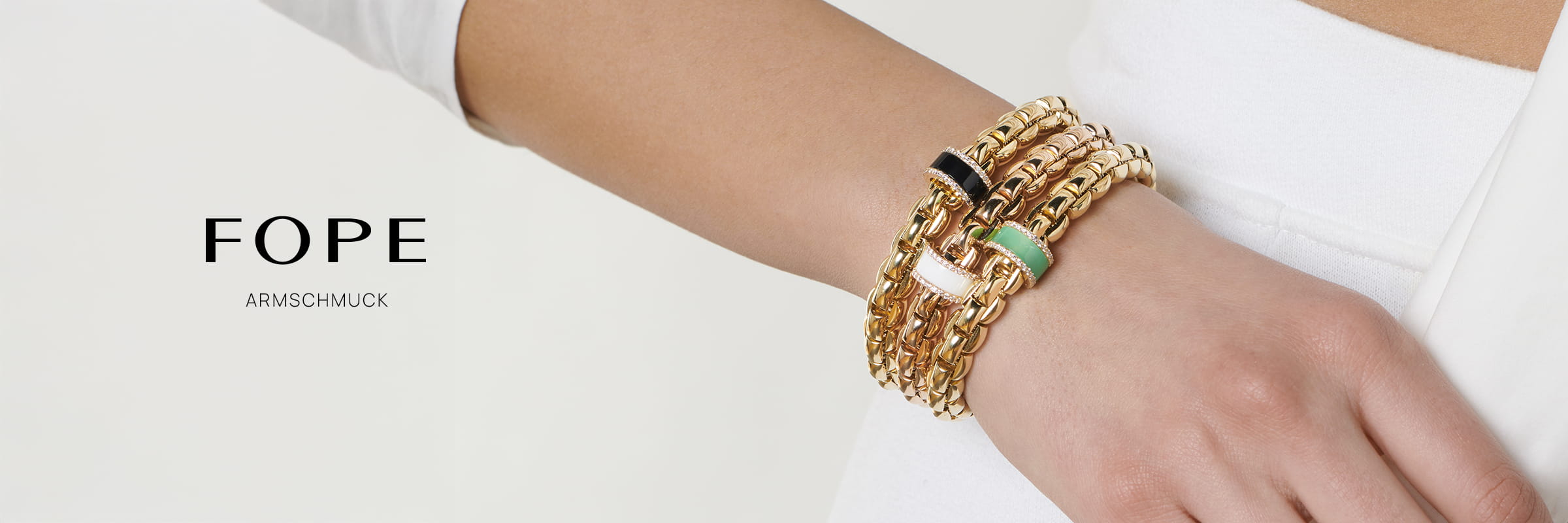 Nahaufnahme eines Handgelenks, das mit Fope Armschmuck geschmückt ist - goldene Kettenarmbänder mit schwarzen, weißen und grünen Akzenten. Auf der linken Seite stehen vor weißem Hintergrund die Worte "FOPE" und "ARMSCHMUCK".