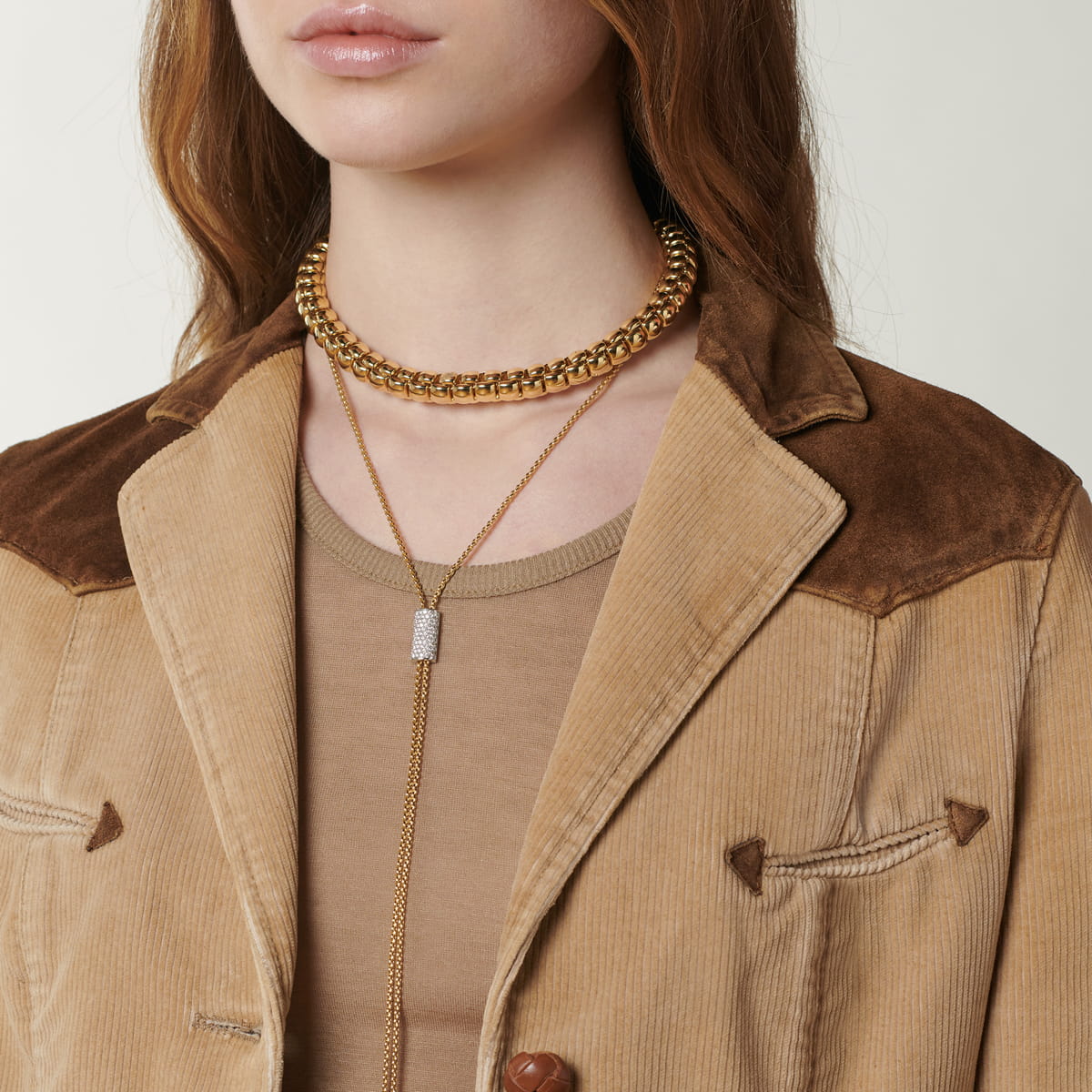 Eine Frau trägt eine hellbraune Cordjacke mit Wildlederdetails, ein beiges Oberteil, eine klobige Goldkette und eine lange goldene Lariat-Halskette mit einem zarten Fope Halsschmuck-Anhänger.