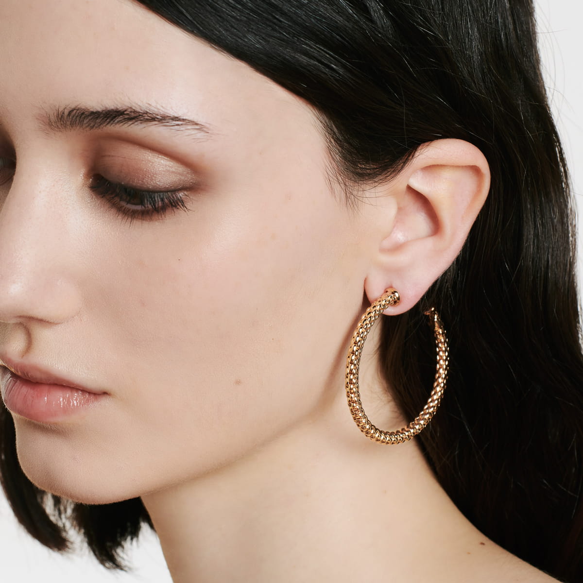 Nahaufnahme einer Person mit heller Haut und dunklem Haar, die einen großen, strukturierten goldenen Fope-Ohrschmuck-Ohrring im linken Ohr trägt und mit neutralem Make-up und ruhigem Gesichtsausdruck nach unten schaut.