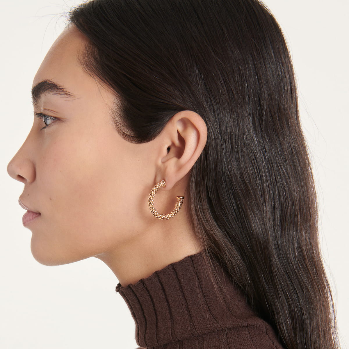 Eine Frau mit langen dunklen Haaren, die einen braunen Rollkragenpullover und einen großen strukturierten Fope-Ohrschmuck-Ohrring aus Gold trägt, ist im Profil vor einem schlichten hellen Hintergrund dargestellt.