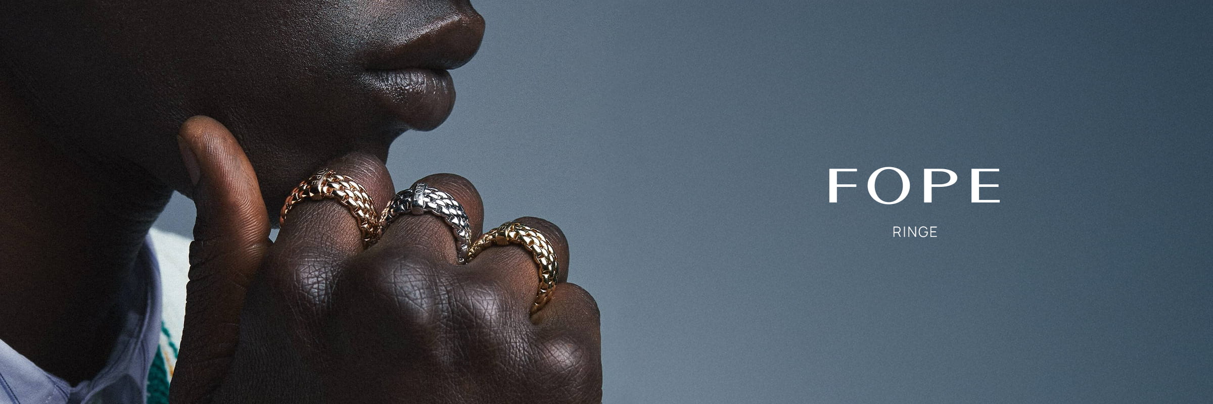 Eine Nahaufnahme der Hand einer Person mit drei strukturierten Fope Ringen in Gold, Silber und Roségold, die sie an ihr Kinn hält. Der Hintergrund ist ein blauer Farbverlauf mit den Worten "FOPE" und "RINGE" auf der rechten Seite.