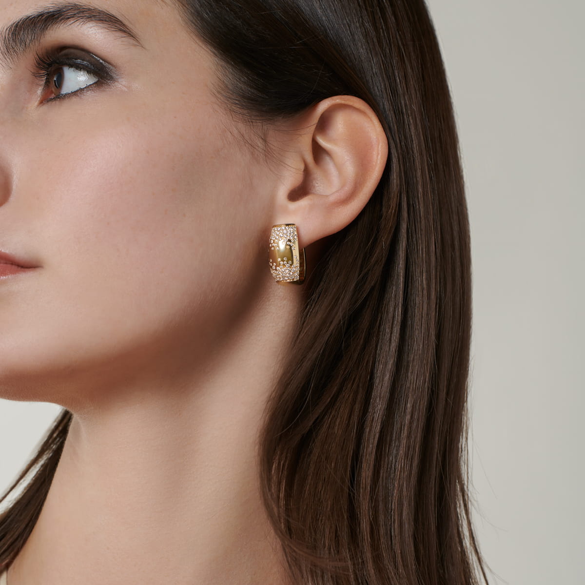 Nahaufnahme von Gesicht und Ohr einer Frau mit glattem braunem Haar und einem großen, rechteckigen Fope-Ohrring aus Gold mit kompliziertem Muster. Der Hintergrund ist neutral und lenkt die Aufmerksamkeit auf das auffällige Schmuckstück.
