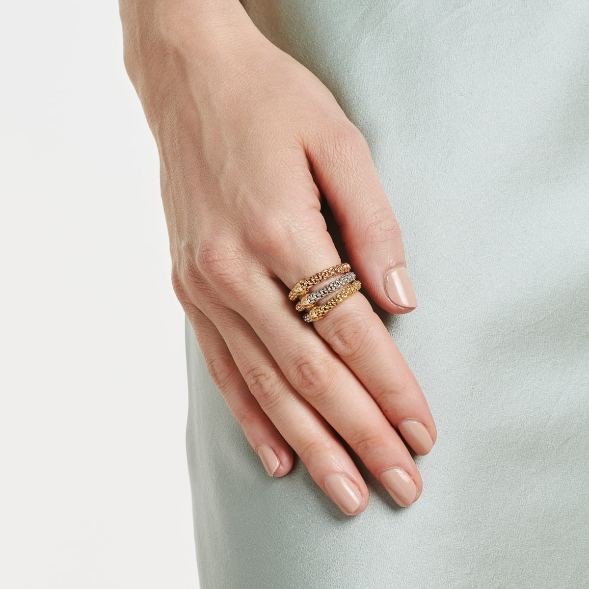 Nahaufnahme einer Hand mit sauber manikürten Nägeln, die auf hellblauem Satinstoff ruht. Die Hand ist mit drei gestapelten Fope-Goldringen geschmückt, die jeweils mit funkelnden Steinen besetzt sind.