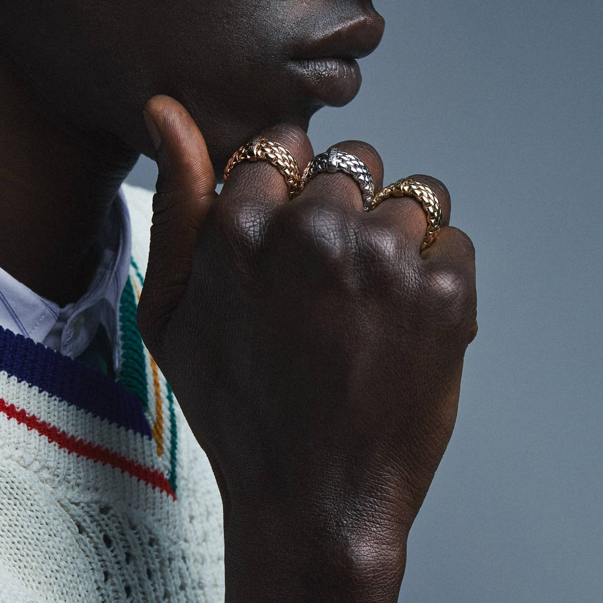 Eine Person mit dunkler Hautfarbe posiert mit ihrer Hand in der Nähe ihres Kinns und trägt drei strukturierte Fope-Ringe in Gold und Silber. Sie trägt einen cremefarbenen Strickpullover mit farbigen Streifen über einem Hemd mit Kragen.