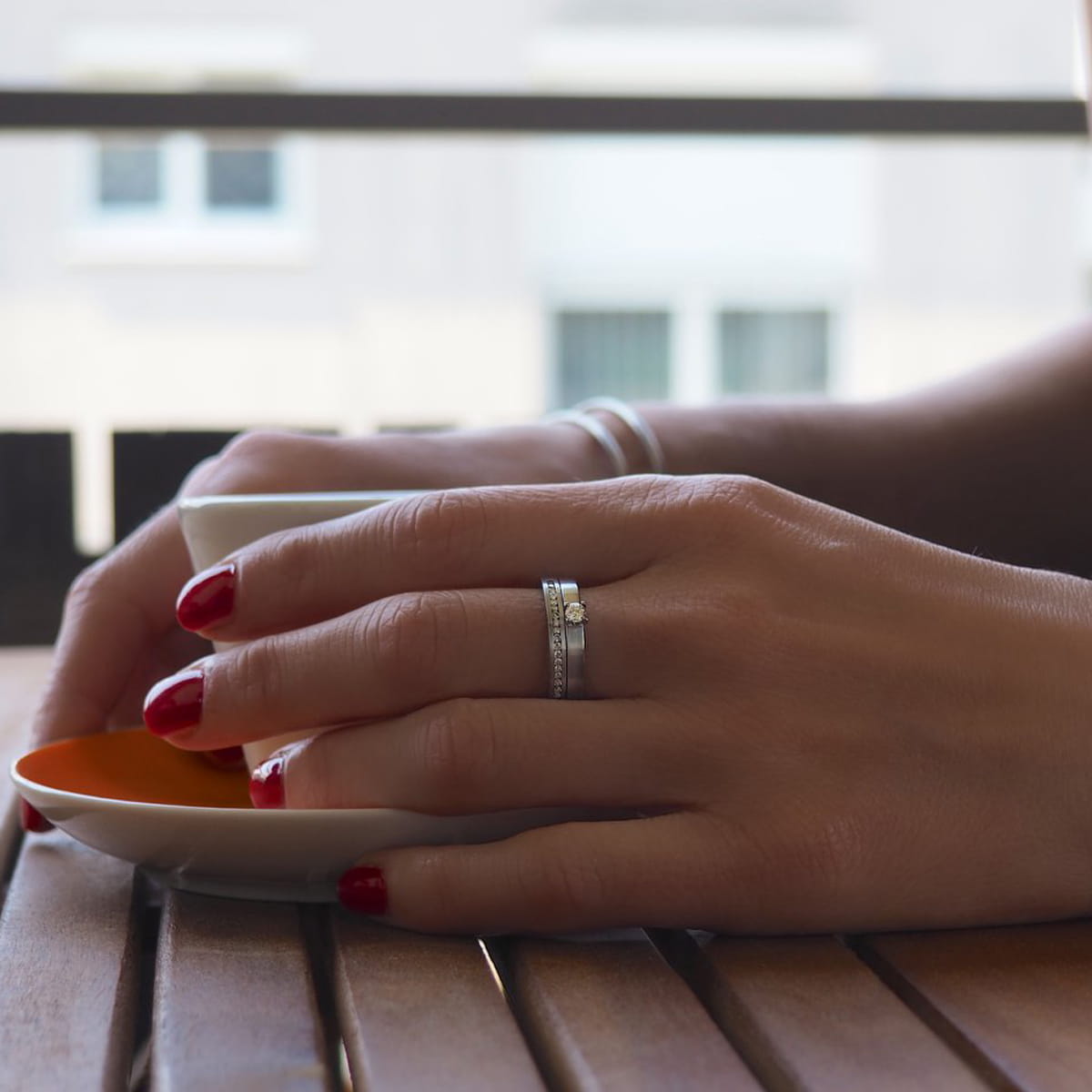 Eine Person mit rot lackierten Nägeln und einem silbernen Ring hält eine weiße Tasse auf einer Untertasse und sitzt an einem Holztisch. Im Hintergrund rahmt ein unscharfes Fenster die gemütliche Szene im Inneren von Humphrey ein.