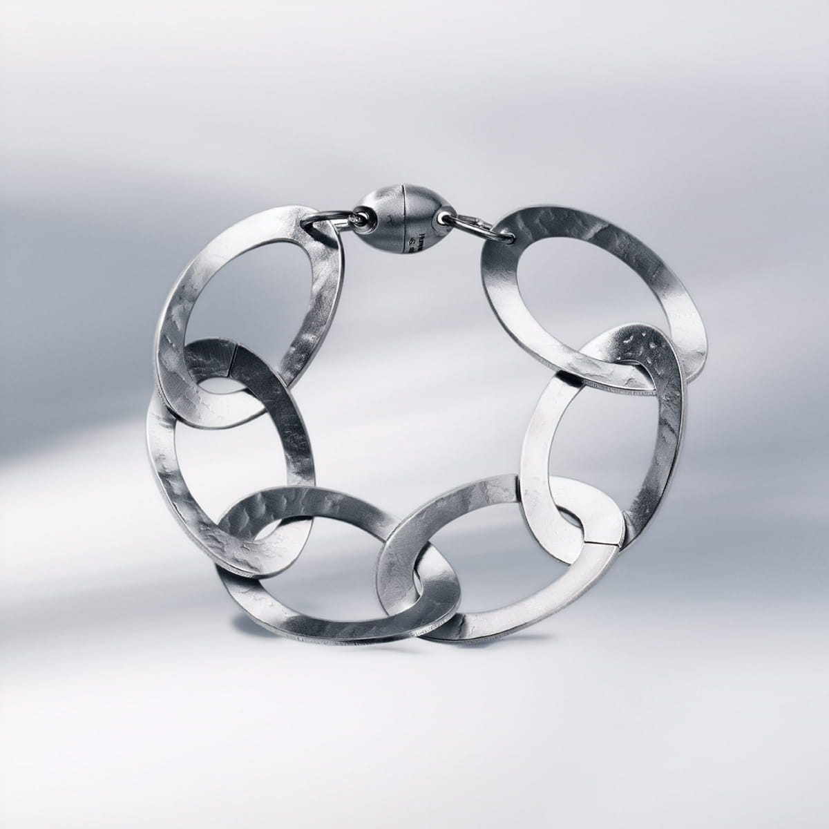 Ein silbernes Humphrey-Armschmuck-Armband mit großen, strukturierten, ovalen Gliedern, die in einer Kette verbunden sind und sich vor einem weichen, hellgrauen Hintergrund präsentieren. Das Armband verfügt über eine runde Schließe an der Spitze.