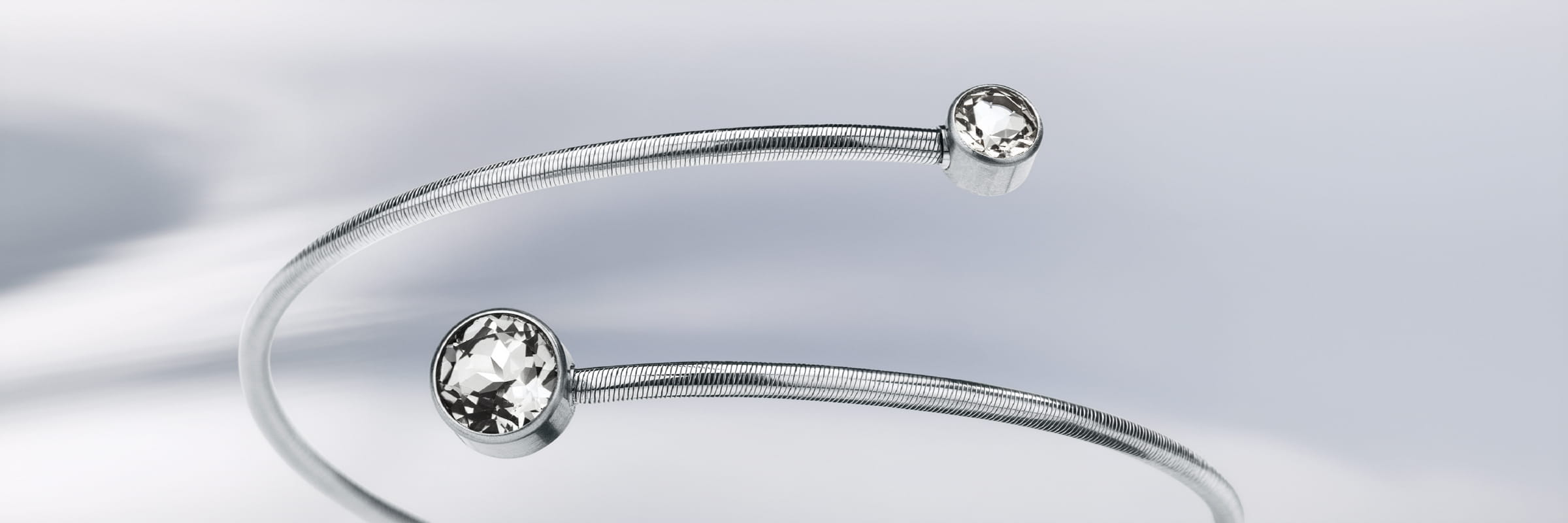 Ein minimalistischer, offener Silberarmreif von Humphrey Armschmuck mit einem runden, klaren Edelstein an jedem Ende, der vor einem weichen, verschwommenen weißen Hintergrund liegt.