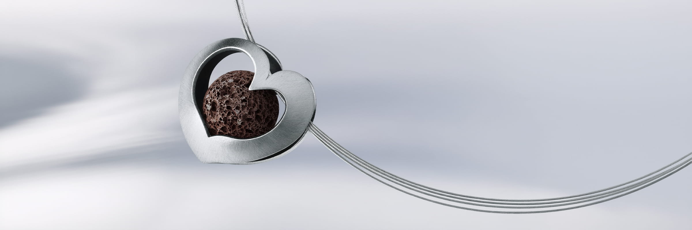 HUMPHREY © 2026 Ein silberner herzförmiger Anhänger mit einem strukturierten dunkelbraunen Stein in der Mitte, Teil der eleganten Humphrey Halsschmuck-Kollektion, hängt an einer dünnen, gebogenen Silberkette vor einem weichen, hellen Hintergrund.