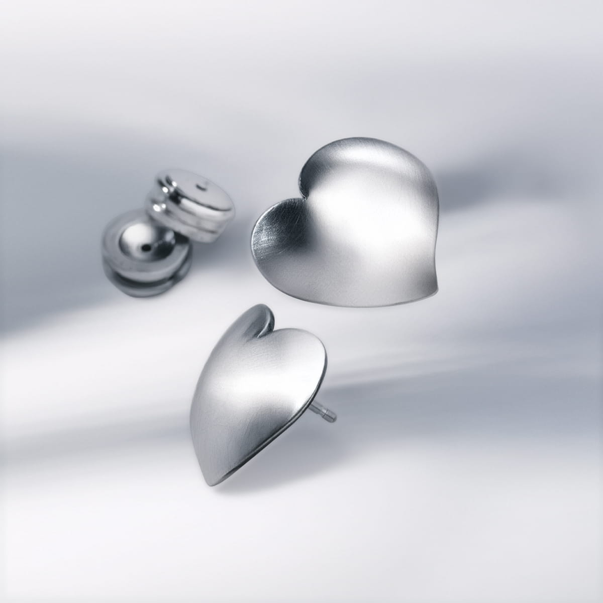 Ein Paar silberner, herzförmiger Ohrstecker von Humphrey Ohrschmuck mit Schmetterlingsverschluss wird auf einer weichen, weißen Oberfläche präsentiert, die sanfte Schatten wirft.