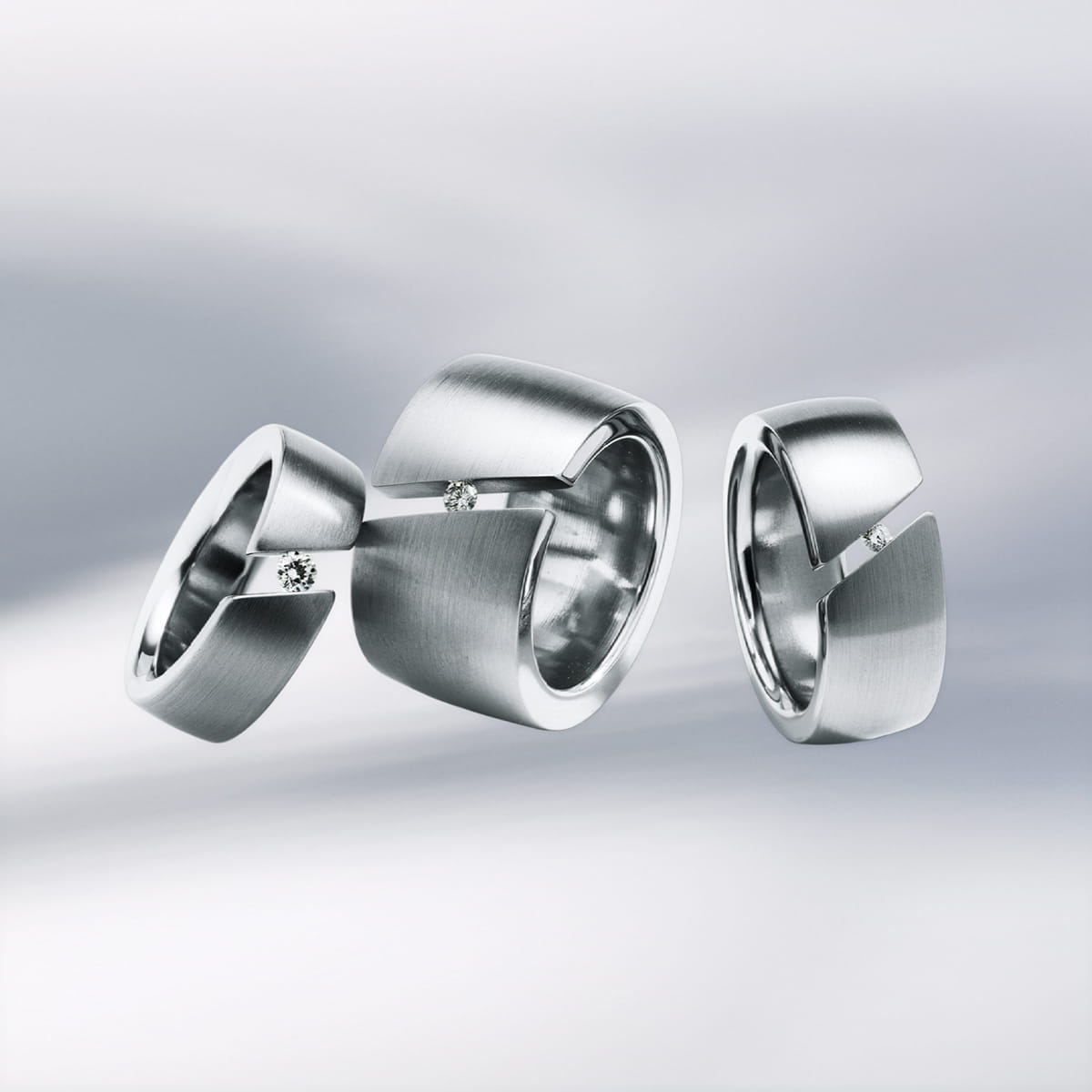 Drei moderne Silberringe von Humphrey Ringe mit matter Oberfläche, die jeweils einen kleinen, in eine diagonale Rille gefassten Diamanten aufweisen, werden vor einem weichen, verschwommenen grauen Hintergrund präsentiert.