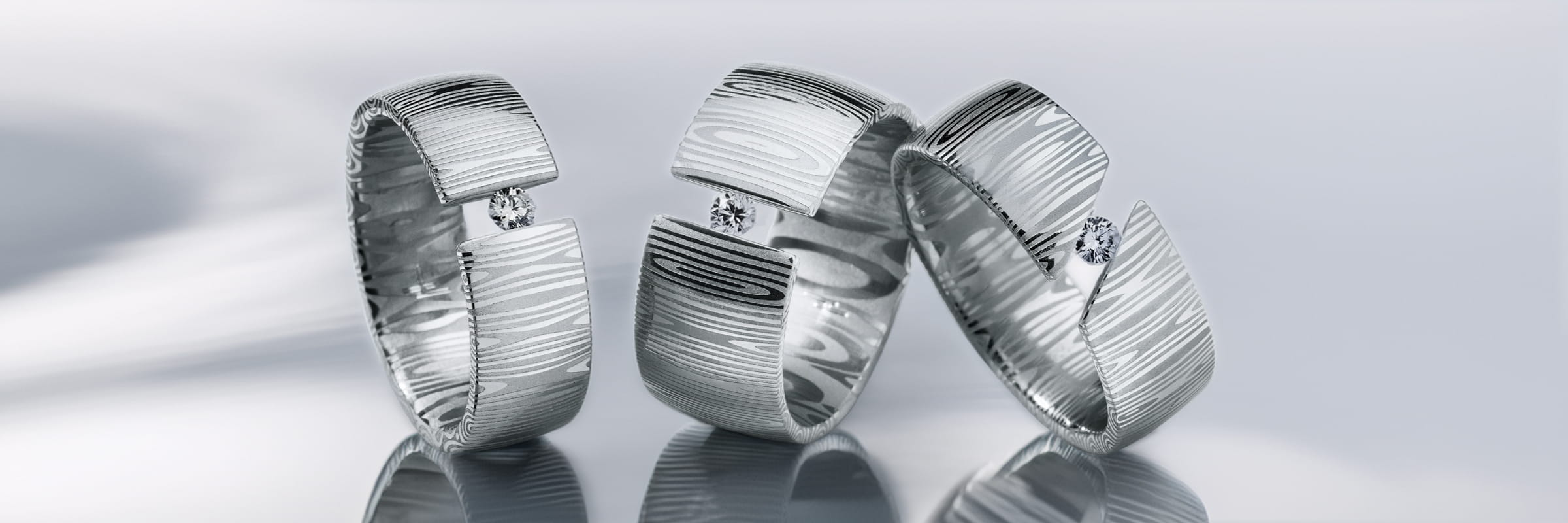 Drei silberne Humphrey Ringe mit einem Holzmaserungsmuster und einem einzelnen runden Diamanten in jedem, aufrecht auf einer reflektierenden Oberfläche vor einem weichen, hellen Hintergrund.