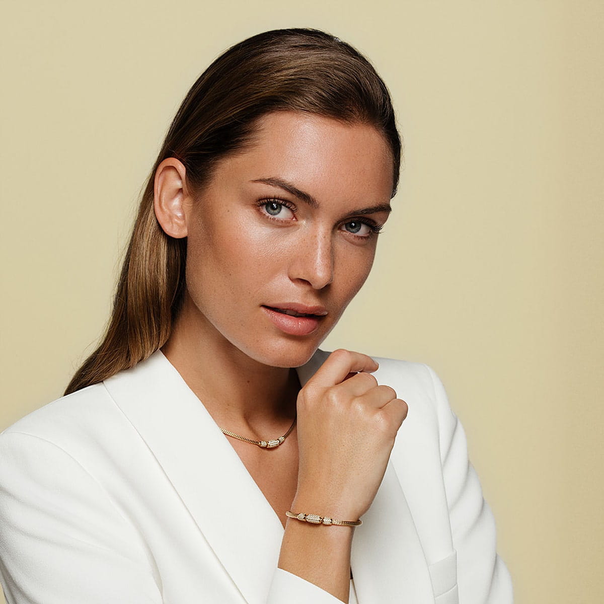 Eine Frau mit hellbraunem Haar trägt einen weißen Blazer, eine goldene Jörg Heinz-Halskette und ein passendes goldenes Armband. Sie blickt mit neutralem Gesichtsausdruck in die Kamera vor einem schlichten beigen Hintergrund.