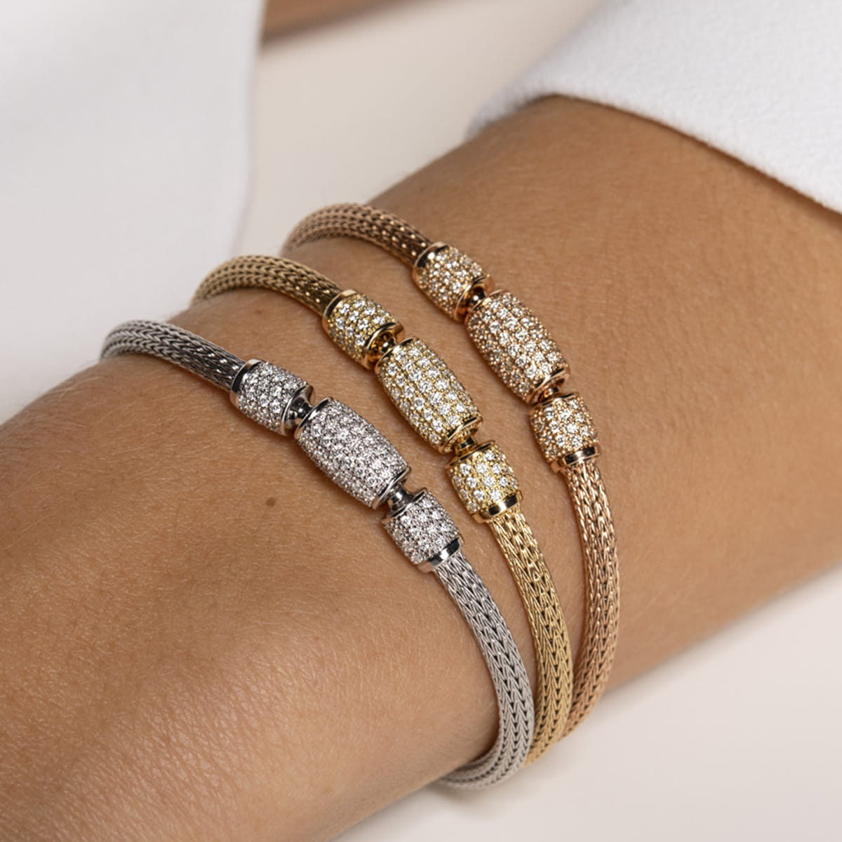 Nahaufnahme eines Handgelenks, das drei Netzarmbänder in Silber-, Gold- und Roségoldtönen trägt, jedes mit einem funkelnden, mit Edelsteinen besetzten zylindrischen Verschluss, der an die Handwerkskunst von Jörg Heinz Halsschmuck erinnert.