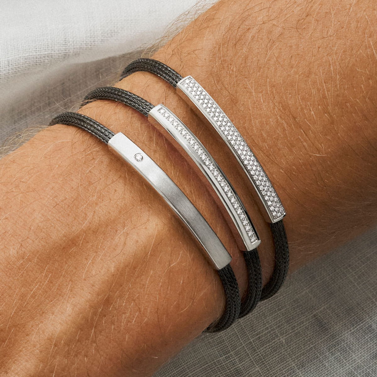 Nahaufnahme eines Handgelenks, das drei schwarze Kordelarmbänder mit je einem silbernen Steg trägt; eines ist schlicht, eines mit einem kleinen Diamanten versehen und eines mit funkelnden Steinen besetzt - ein Hinweis auf die raffinierte Eleganz von Jörg Heinz Halsschmuck.