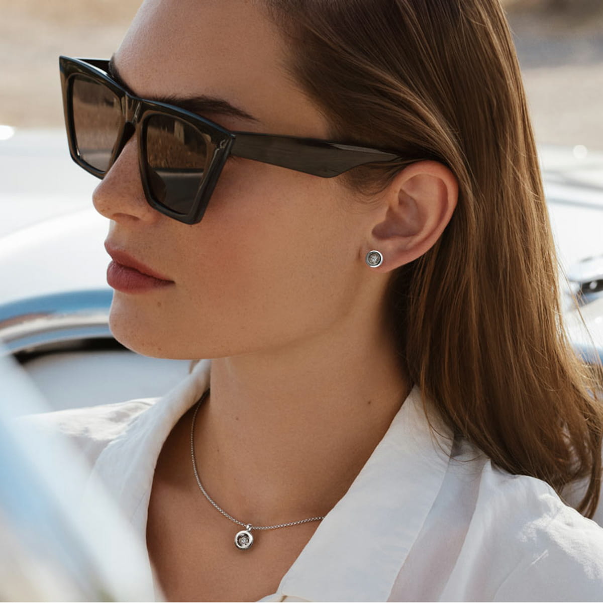 Eine Frau mit großer schwarzer Sonnenbrille, Ohrsteckern und einer passenden Halskette mit Jörg Heinz Halsschmuck-Anhänger sitzt in einem weißen Hemd, das Sonnenlicht beleuchtet ihr Gesicht und der Hintergrund im Freien ist unscharf.