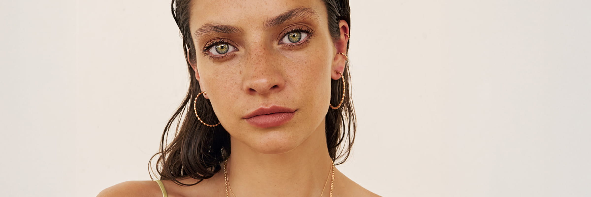 Eine Frau mit nassen dunklen Haaren, grünen Augen und Sommersprossen starrt in die Kamera. Sie trägt große Jörg Heinz Ohrschmuck-Ohrringe, mehrlagige Halsketten und ein helles Oberteil vor einem schlichten, hellen Hintergrund.