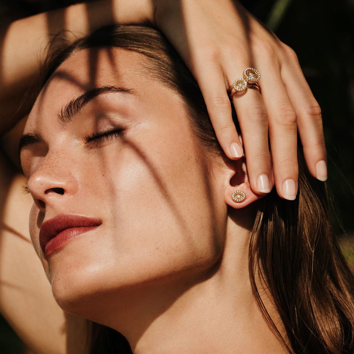 Eine Frau mit geschlossenen Augen stützt ihren Kopf auf ihre Hand. Sie trägt goldene Ohrringe und Jörg Heinz Ringe. Das Sonnenlicht fällt durch die Blätter und wirft Schatten auf ihr Gesicht und ihre Hand.