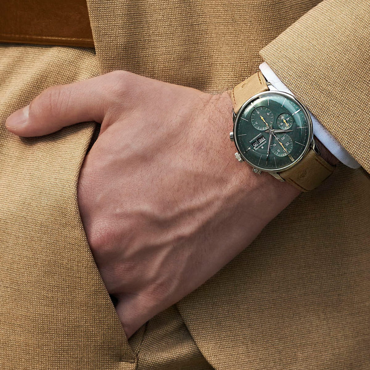 JUNGHANS © 2026 Eine Person, die einen hellbraunen Anzug trägt, hat die linke Hand in der Tasche und zeigt eine stilvolle Junghans-Armbanduhr mit grünem Zifferblatt und hellbraunem Lederarmband.