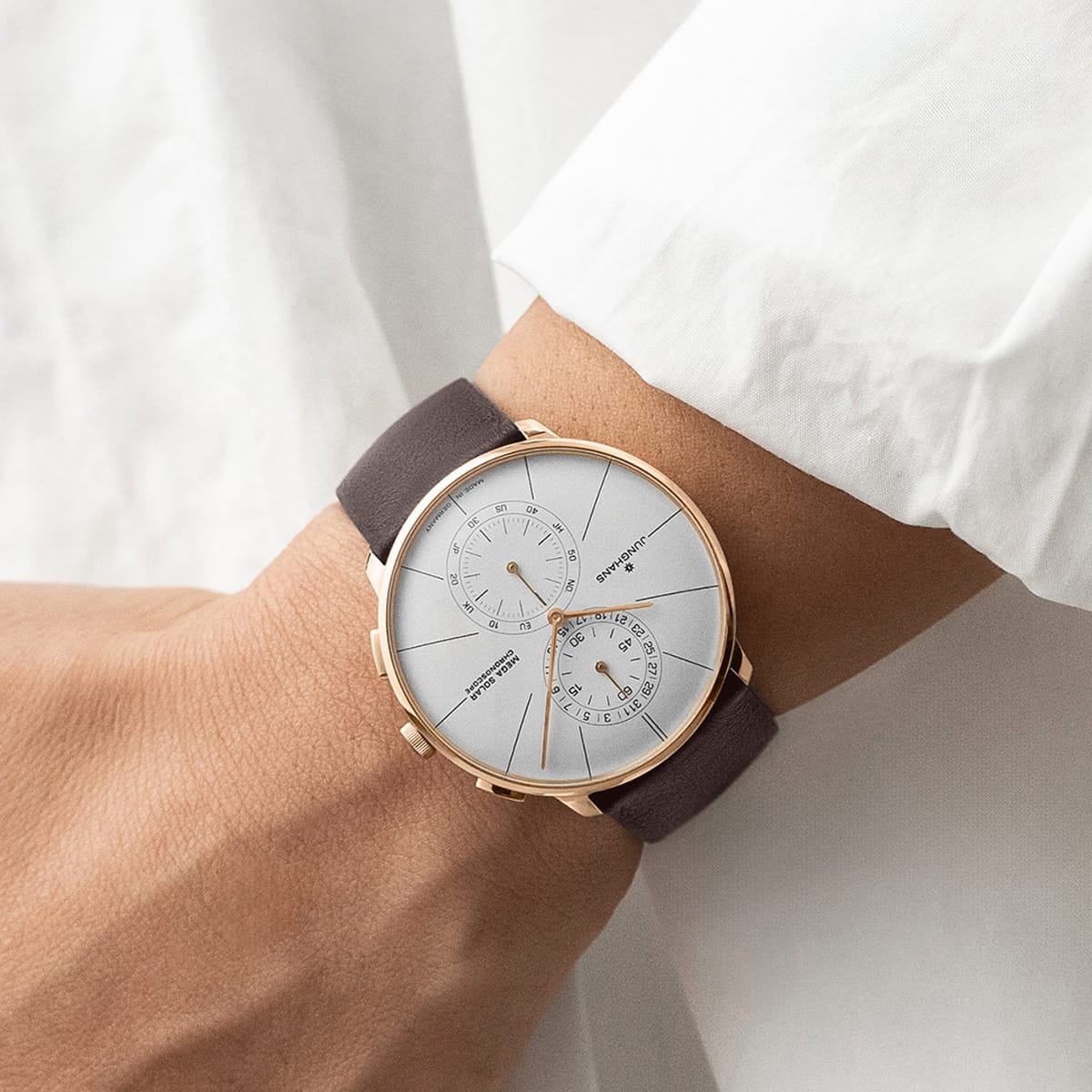 JUNGHANS © 2026 Eine Person, die ein weißes, langärmeliges Hemd trägt, wird mit einer stilvollen Junghans-Armbanduhr mit rundem, weißem Zifferblatt, goldener Lünette, zwei Hilfszifferblättern und einem dunkelbraunen Lederarmband gezeigt.