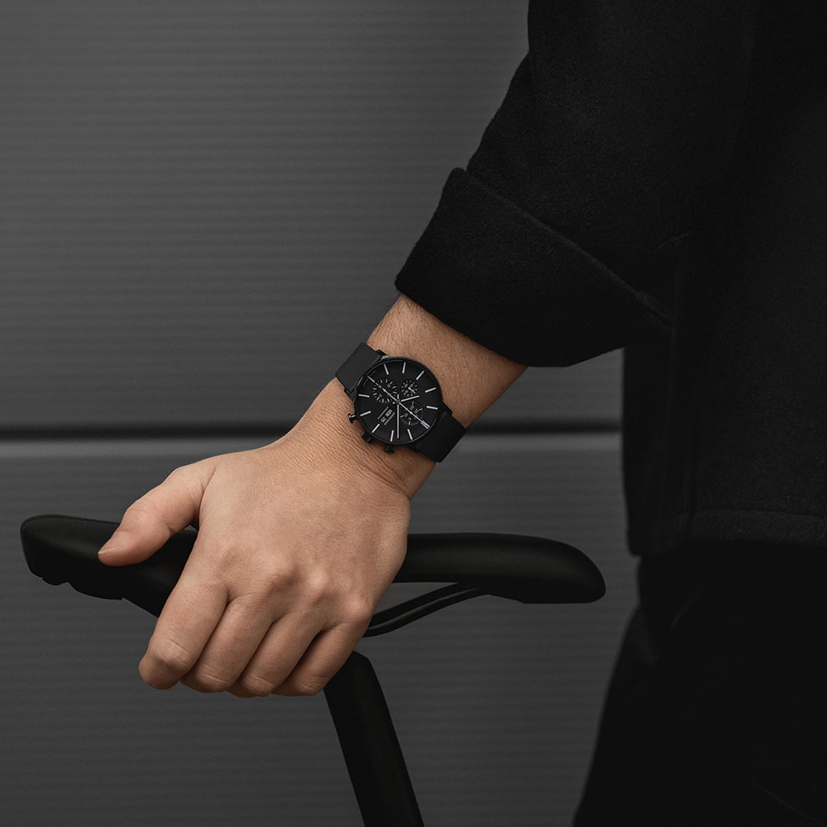 JUNGHANS © 2026 Eine Person, die eine schwarze Junghans-Uhr trägt, stützt ihre Hand auf den Sitz eines Fahrrads. Der Hintergrund ist unscharf und grau, und die Person ist schwarz gekleidet.