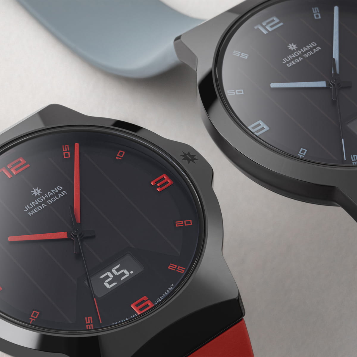 JUNGHANS © 2026 Nahaufnahme von zwei Junghans Mega Solar Armbanduhren mit schwarzem Zifferblatt - eine mit roten Stundenmarkierungen und digitaler Datumsanzeige, die andere blau. Beide Junghans-Uhren haben ein Metallgehäuse und werden auf einer hellen Fläche präsentiert.