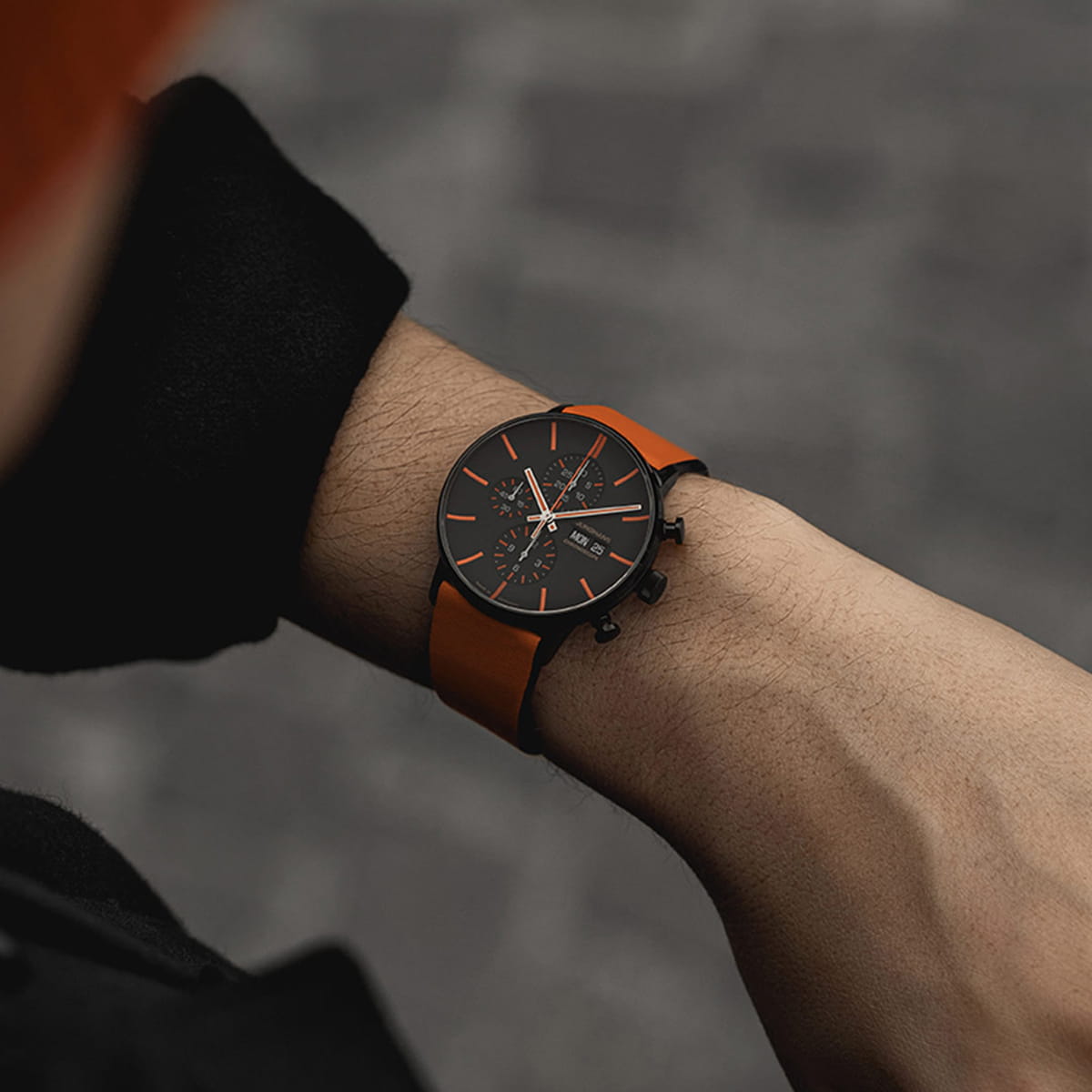 JUNGHANS © 2026 Eine Person trägt eine schwarze Junghans-Form-Uhr mit orangefarbenem Armband und orangefarbenen Details auf dem Zifferblatt, die in Nahaufnahme am Handgelenk vor einem unscharfen grauen Hintergrund gezeigt wird.