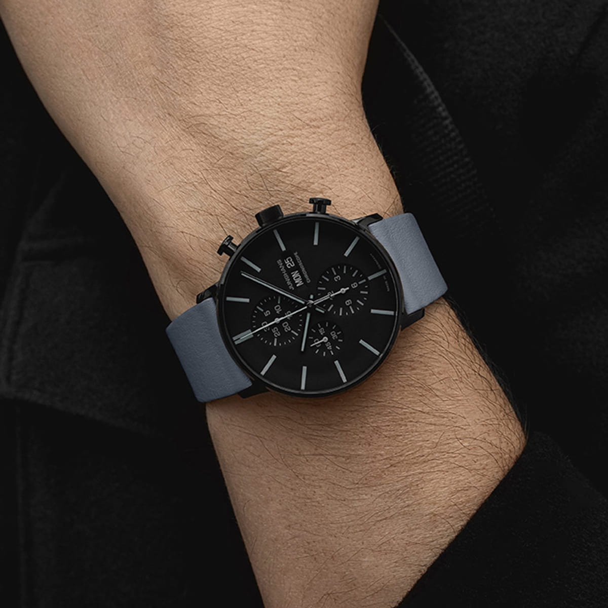 JUNGHANS © 2026 Eine Person trägt eine schwarze Junghans-Form-Chronographen-Armbanduhr mit einem dunkelgrauen Lederarmband und einem schwarzen Zifferblatt mit drei Hilfszifferblättern und weißen Stundenmarkierungen. Die Uhr befindet sich am linken Handgelenk, und die Person ist schwarz gekleidet.