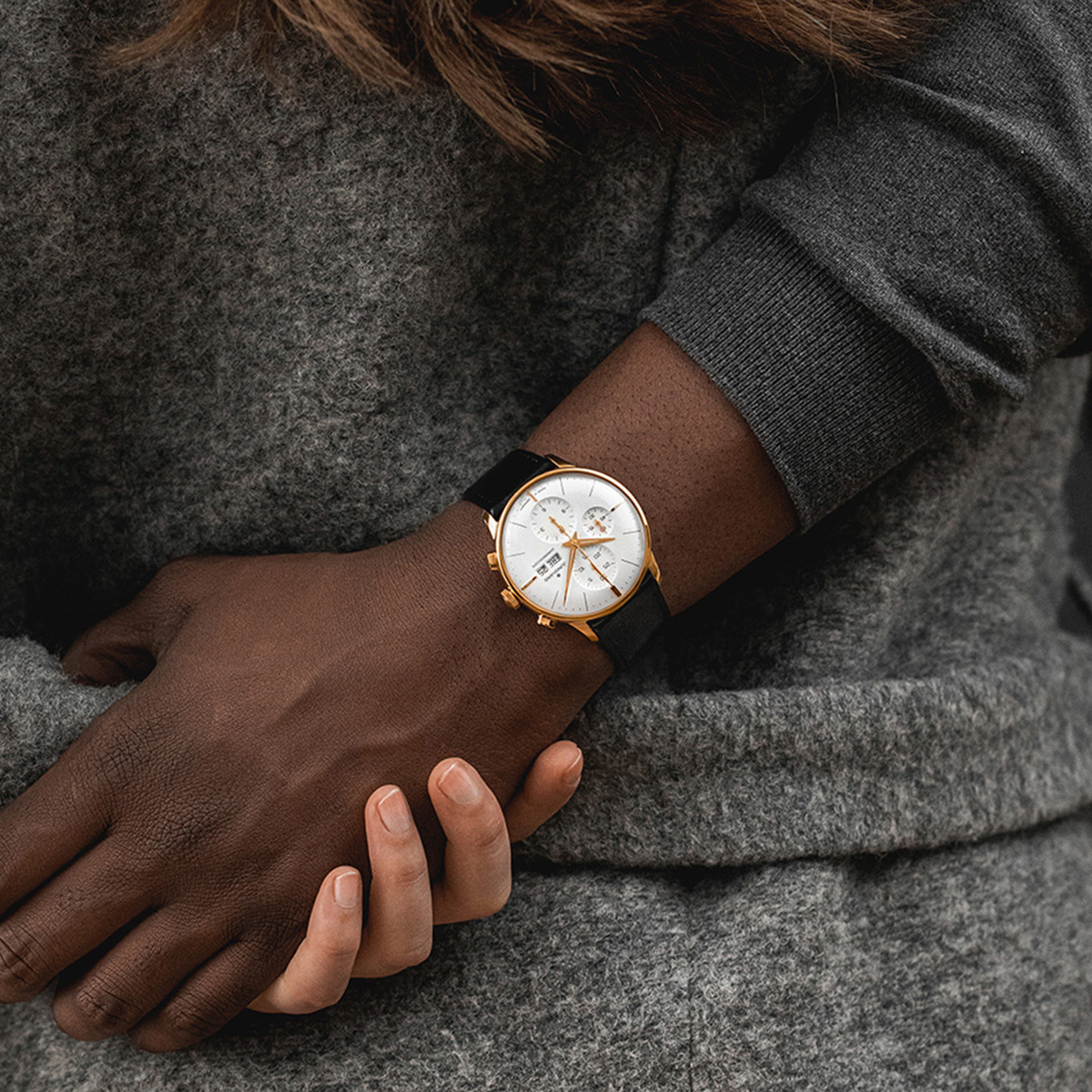 Eine Person, die einen grauen Pullover trägt, verschränkt die Arme und präsentiert eine goldene Junghans Meister Kollektion Armbanduhr mit weißem Zifferblatt und schwarzem Armband an ihrem linken Handgelenk. Ihre Fingernägel sind ordentlich geschnitten und die Haare sind teilweise sichtbar.