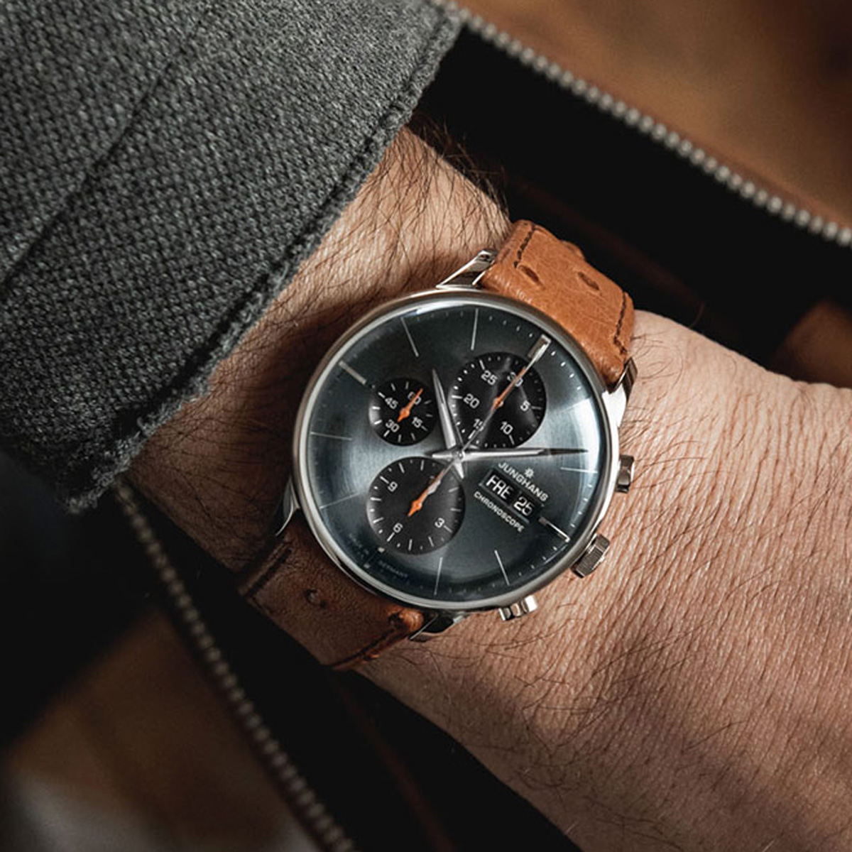 Eine Person, die eine graue Jacke trägt, zeigt eine Junghans Meister Kollektion Armbanduhr mit einem braunen Lederarmband und einem großen, runden schwarzen Zifferblatt mit mehreren Hilfszifferblättern und silbernen Stundenmarkierungen.