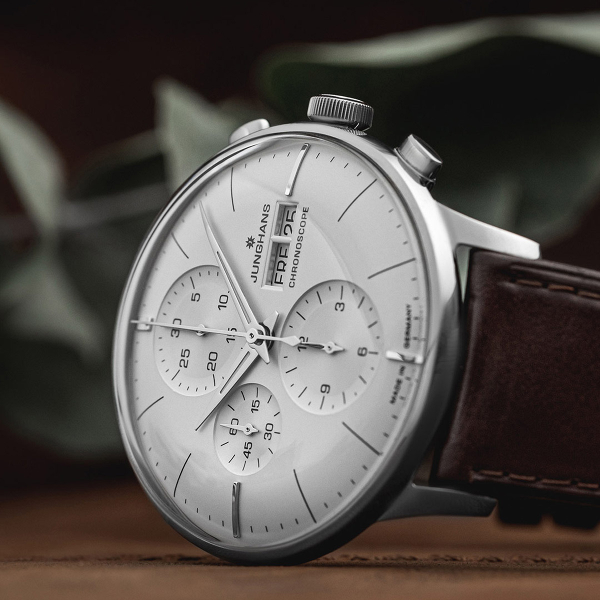 Eine Nahaufnahme einer silbernen Junghans Meister Kollektion Chronographen-Armbanduhr mit weißem Zifferblatt, mehreren Hilfszifferblättern und braunem Lederarmband vor einem unscharfen Hintergrund mit grünen Blättern.
