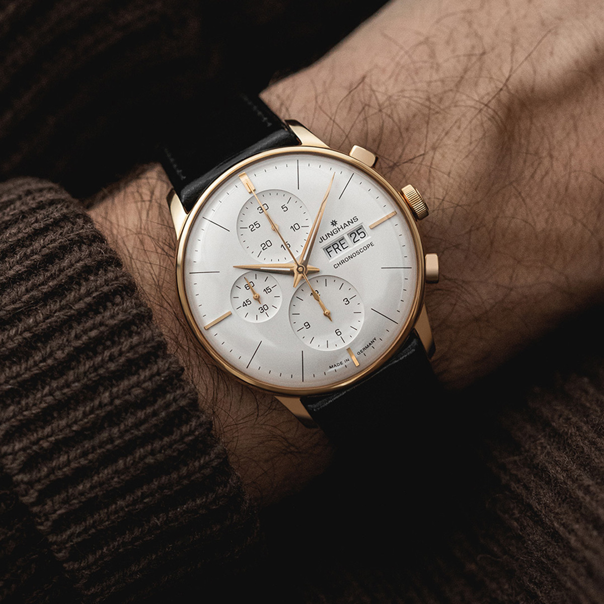 Nahaufnahme einer Junghans Meister Kollektion Armbanduhr mit Goldgehäuse, weißem Zifferblatt, Chronographenfunktion und schwarzem Lederarmband, getragen an einem Handgelenk in einem braunen Strickpullover. Die Uhr zeigt den Tag und das Datum an.